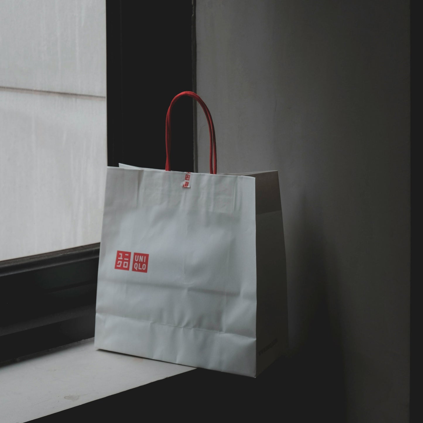 White paper bag with red handles and logo leaning against a wall.