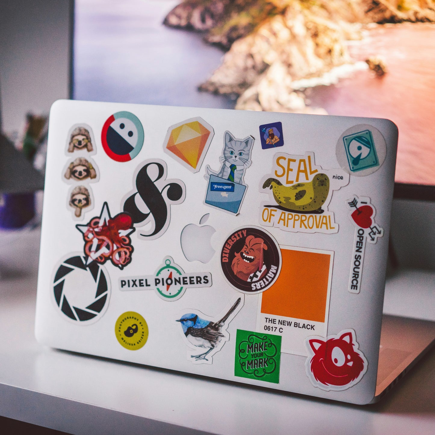 Laptop with various stickers on a desk with computer monitors in the background